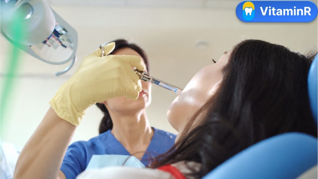 Bàn tay đeo găng đưa ống tiêm đến vùng miệng người bệnh trên ghế nha khoa